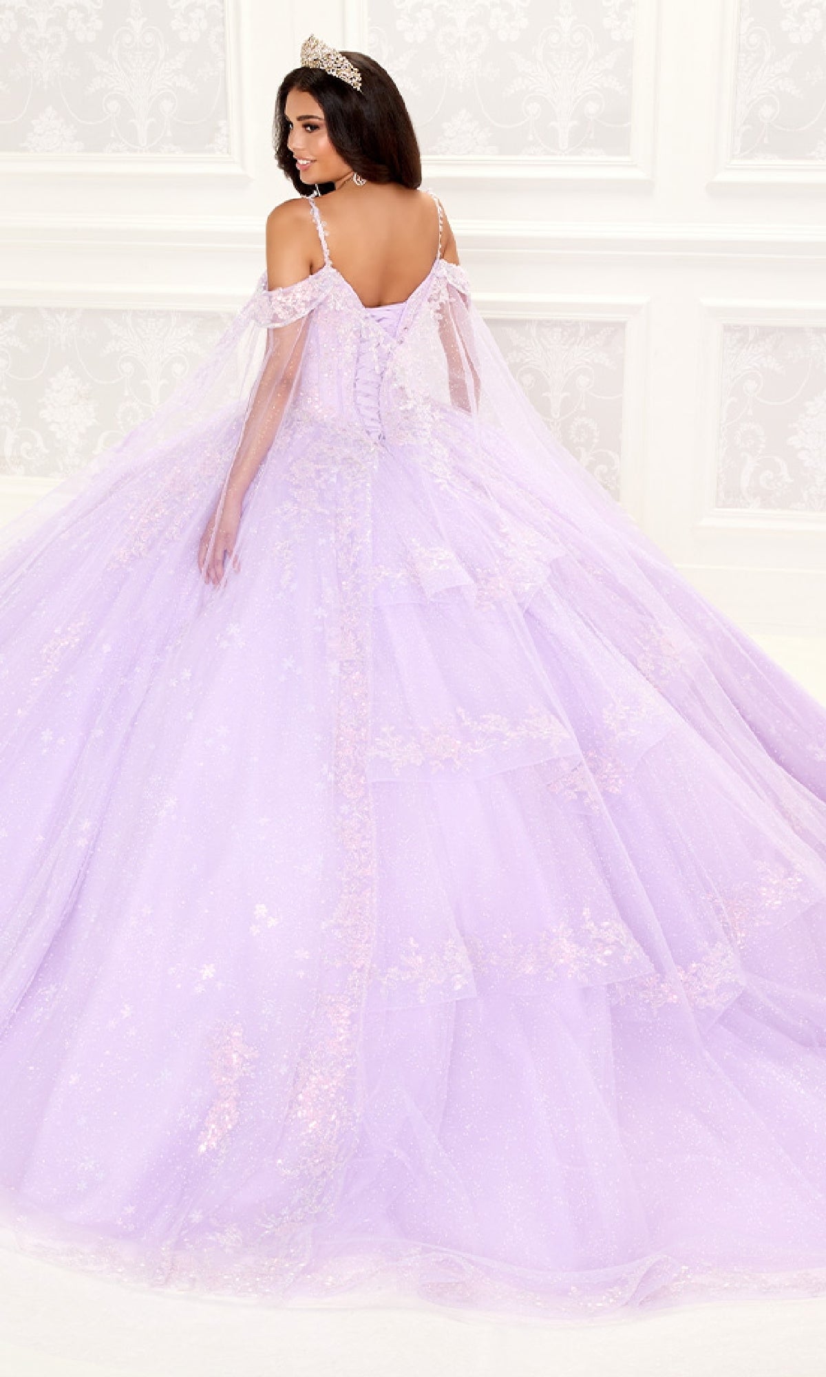A woman in the Quinceanera Dress PR30087 By Princesa, featuring sparkling lavender fabric, off-the-shoulder sleeves, and a corset bodice with a tiara, stands before an ornate white wall, facing away but glancing over her shoulder.